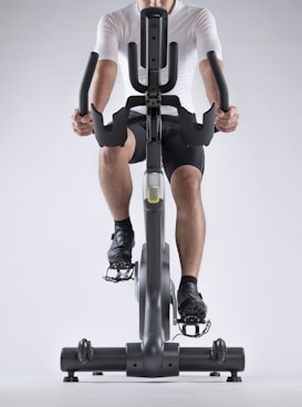A person dressed in athletic gear is actively pedaling on a stationary exercise bike, with a focus on their lower body and cycling position. The background is minimalistic, highlighting the exercise equipment and individual.