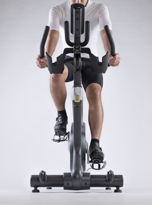A person dressed in athletic gear is actively pedaling on a stationary exercise bike, with a focus on their lower body and cycling position. The background is minimalistic, highlighting the exercise equipment and individual.