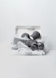 A white gift box contains two grey caps, wrapped items with visible text, and rolled cloths. The items have an organized and neat arrangement, creating a minimalist aesthetic.