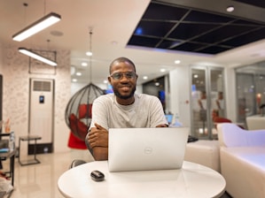 A man is sitting at a round white table using a laptop. He is indoors in a modern office setting with a hanging chair and contemporary lighting. The background includes a glass partition and soft furnishings.