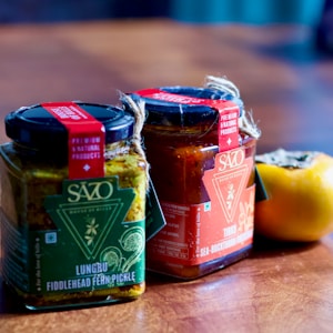 Two jars filled with pickles are placed on a wooden surface. One jar contains fiddlehead fern pickle with a green label, and the other has sea-buckthorn pickle with a red label. Both jars have lids secured with twine, along with branding and product information. A persimmon lies next to the jars, adding a touch of natural, vibrant color.