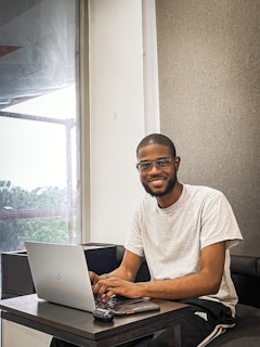 A person is sitting at a desk working on a laptop, with a smile on their face. The desk also has a mouse and a phone placed on it. The individual is wearing a light gray t-shirt and black pants, and they are in a room with a large window that shows some trees outside.