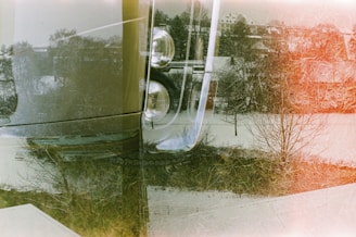 A double exposure photograph combining elements of a snowy landscape and a close-up of a vehicle's headlight. Trees without leaves and rooftops dusted with snow are visible in the background, while the curved lines and reflective surfaces of the car's headlamp dominate the foreground.