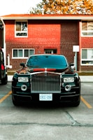 A luxury car parked in front of a brick residential building. The car is black with a prominent grille and distinctive headlights. It is positioned between parking lines on a paved area. The building in the background has multiple windows and a slightly overhanging roof. There are trees partially visible above the building.