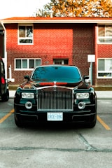 A luxury car parked in front of a brick residential building. The car is black with a prominent grille and distinctive headlights. It is positioned between parking lines on a paved area. The building in the background has multiple windows and a slightly overhanging roof. There are trees partially visible above the building.