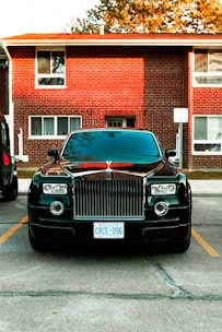 A luxury car parked in front of a brick residential building. The car is black with a prominent grille and distinctive headlights. It is positioned between parking lines on a paved area. The building in the background has multiple windows and a slightly overhanging roof. There are trees partially visible above the building.