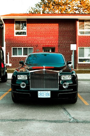 A luxury car parked in front of a brick residential building. The car is black with a prominent grille and distinctive headlights. It is positioned between parking lines on a paved area. The building in the background has multiple windows and a slightly overhanging roof. There are trees partially visible above the building.