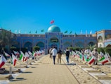 A courtyard with rows of graves, each adorned with an Iranian flag. People are walking through the area, which is surrounded by buildings with intricate blue tile work. Green banners are also displayed above the building, and a blue dome is visible in the center.