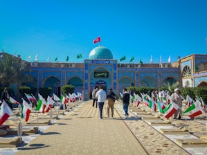 A courtyard with rows of graves, each adorned with an Iranian flag. People are walking through the area, which is surrounded by buildings with intricate blue tile work. Green banners are also displayed above the building, and a blue dome is visible in the center.