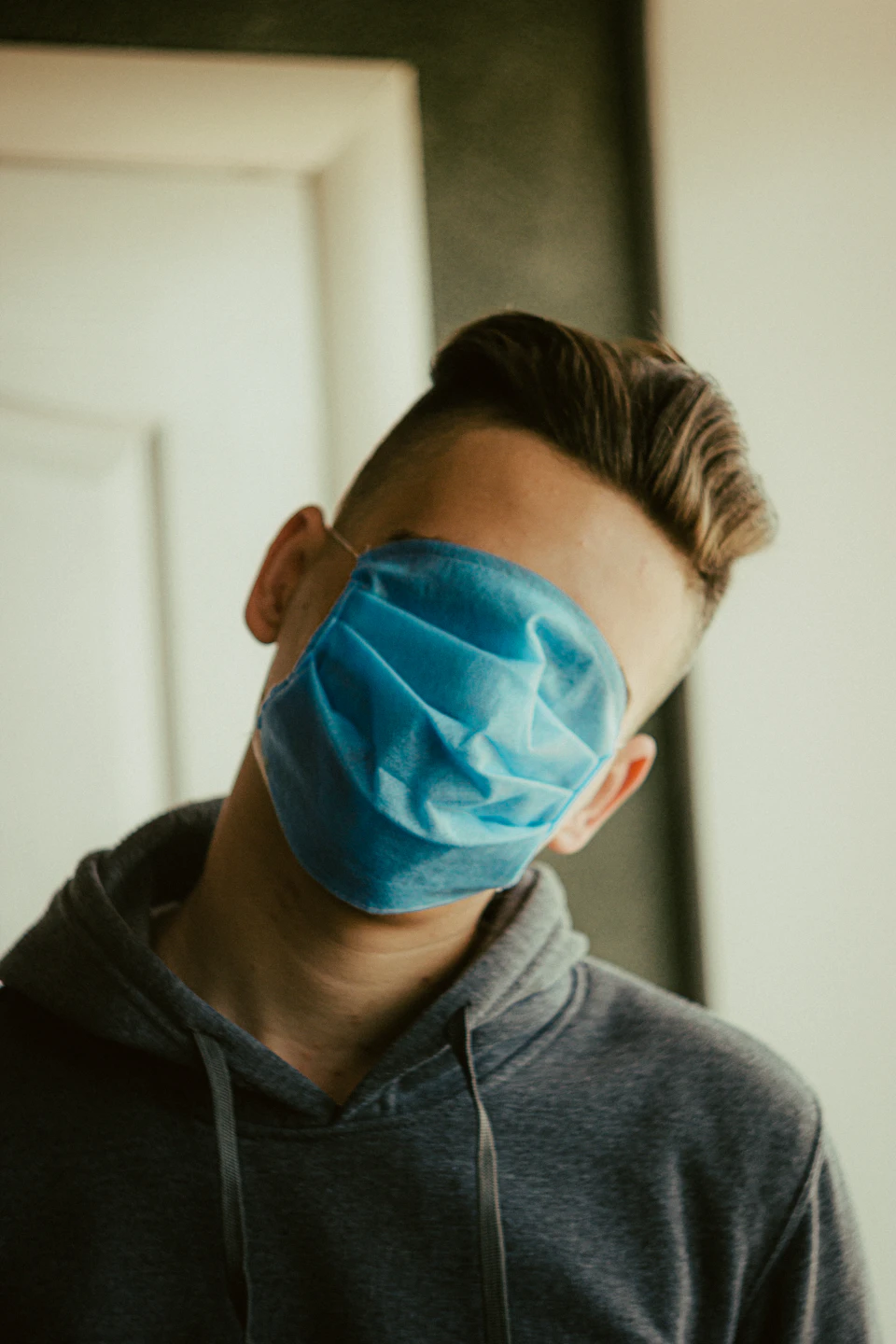 In the image, a young man is captured wearing a blue surgical mask over his eyes and nose. He is standing against a white door backdrop, which contrasts with his dark hair and gray hoodie. The mask covers most of his face, obscuring his features from view.