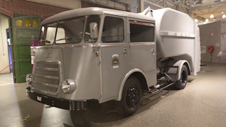 A vintage utility vehicle with a grey metallic finish is displayed indoors. It has a rounded cabin with large, curved windows and a prominent front grille. The body design is mid-20th century, with a large storage or transport compartment in the back. The wheels are black, and the vehicle features a small crest or emblem on the side. The background includes brick walls and industrial elements, suggesting a museum or exhibit setting.