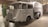 A vintage utility vehicle with a grey metallic finish is displayed indoors. It has a rounded cabin with large, curved windows and a prominent front grille. The body design is mid-20th century, with a large storage or transport compartment in the back. The wheels are black, and the vehicle features a small crest or emblem on the side. The background includes brick walls and industrial elements, suggesting a museum or exhibit setting.