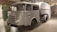 A vintage utility vehicle with a grey metallic finish is displayed indoors. It has a rounded cabin with large, curved windows and a prominent front grille. The body design is mid-20th century, with a large storage or transport compartment in the back. The wheels are black, and the vehicle features a small crest or emblem on the side. The background includes brick walls and industrial elements, suggesting a museum or exhibit setting.