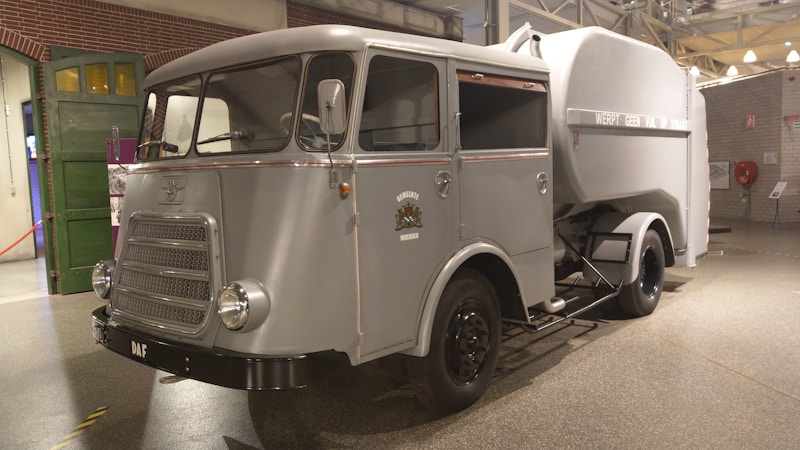 A vintage utility vehicle with a grey metallic finish is displayed indoors. It has a rounded cabin with large, curved windows and a prominent front grille. The body design is mid-20th century, with a large storage or transport compartment in the back. The wheels are black, and the vehicle features a small crest or emblem on the side. The background includes brick walls and industrial elements, suggesting a museum or exhibit setting.