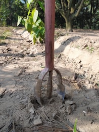 A gardening tool with a long handle is embedded in dry, dusty soil. The visible part of the tool has several prongs, indicating it's used for digging or cultivating. Surrounding the tool are scattered dried leaves and a small green plant to the left. In the background, there are some trees providing partial shade.