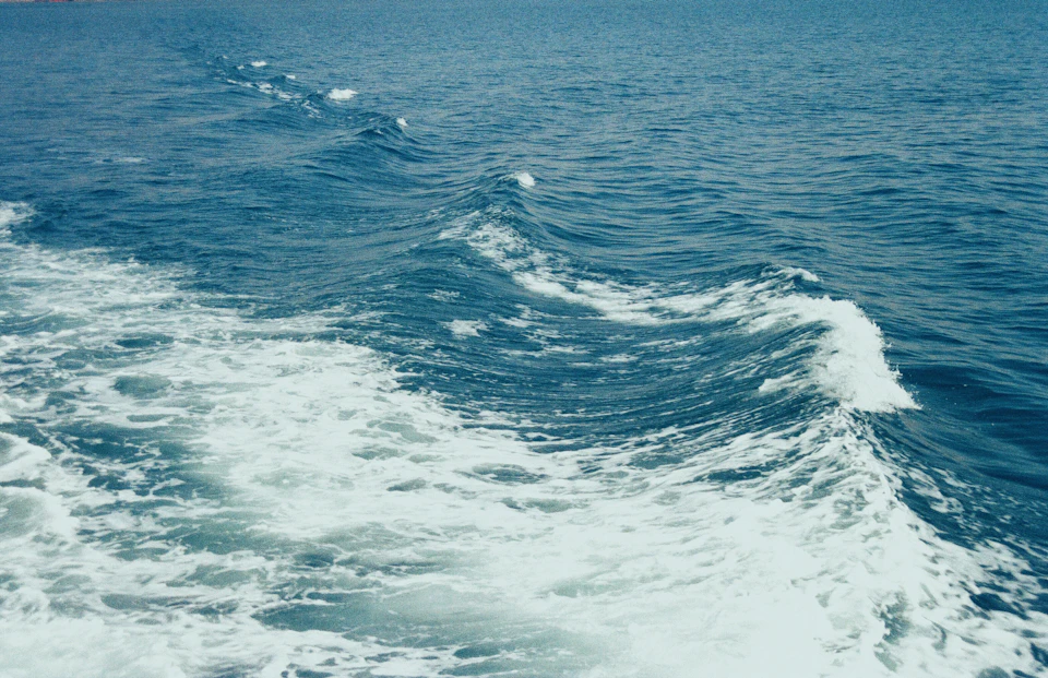 The image depicts a serene ocean scene with a large wave breaking towards the right side, creating a beautiful contrast against the deep blue water. The wave is white and frothy on top, indicating its power and movement. The horizon line can be seen in the background, adding depth to the image. The sky above mirrors the color of the sea below, blending seamlessly into the scene.