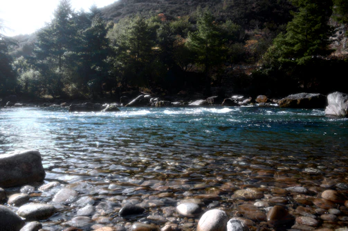A clear river flowing gently over smooth stones under a bright sky.