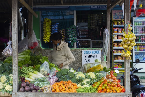 A person stands at a vegetable stall that is filled with a variety of fresh vegetables, including carrots, tomatoes, broccoli, cauliflower, eggplants, and green beans. The person appears to be arranging or handling the produce. In the background, there are shelves stocked with various packaged goods and snacks in a small market setup. There is a signboard that reads 'GREEN HOUSE SIMPATI' and some hanging garlic on the right side of the stall.