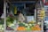 A person stands at a vegetable stall that is filled with a variety of fresh vegetables, including carrots, tomatoes, broccoli, cauliflower, eggplants, and green beans. The person appears to be arranging or handling the produce. In the background, there are shelves stocked with various packaged goods and snacks in a small market setup. There is a signboard that reads 'GREEN HOUSE SIMPATI' and some hanging garlic on the right side of the stall.
