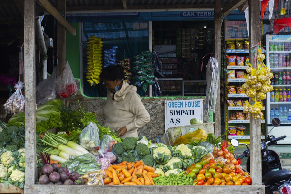 A person stands at a vegetable stall that is filled with a variety of fresh vegetables, including carrots, tomatoes, broccoli, cauliflower, eggplants, and green beans. The person appears to be arranging or handling the produce. In the background, there are shelves stocked with various packaged goods and snacks in a small market setup. There is a signboard that reads 'GREEN HOUSE SIMPATI' and some hanging garlic on the right side of the stall.