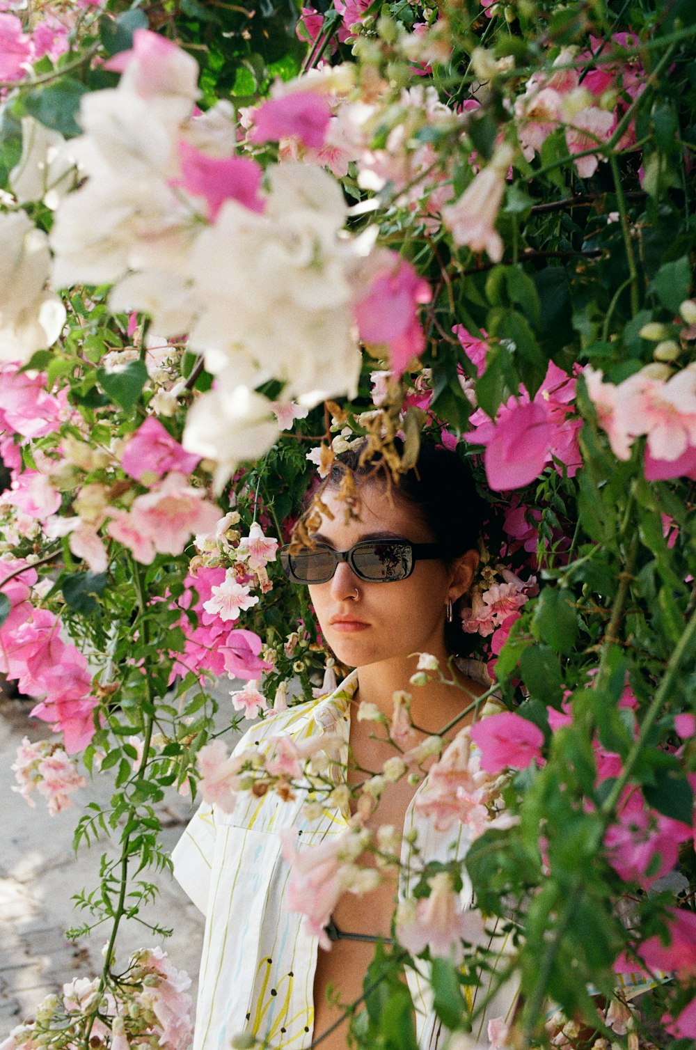 Una mujer con gafas de sol parada bajo un arbusto de flores rosadas y blancas