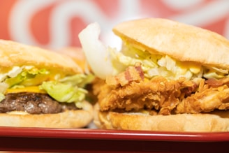 Two sandwiches are placed side by side on a red tray. One sandwich contains a beef patty with lettuce and cheese, while the other features fried chicken with bacon and lettuce. The bread appears soft and slightly toasted. The background features a blurred red and white pattern.