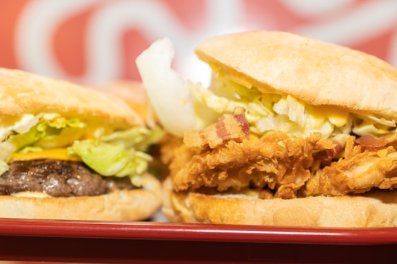 Two sandwiches are placed side by side on a red tray. One sandwich contains a beef patty with lettuce and cheese, while the other features fried chicken with bacon and lettuce. The bread appears soft and slightly toasted. The background features a blurred red and white pattern.