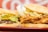 Two sandwiches are placed side by side on a red tray. One sandwich contains a beef patty with lettuce and cheese, while the other features fried chicken with bacon and lettuce. The bread appears soft and slightly toasted. The background features a blurred red and white pattern.