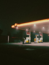 A dimly lit gas station at night, with a retro aesthetic. The scene is characterized by a glowing canopy with an orange tint, three gas pumps beneath it, and a backdrop of darkness surrounding the area.
