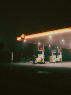 A dimly lit gas station at night, with a retro aesthetic. The scene is characterized by a glowing canopy with an orange tint, three gas pumps beneath it, and a backdrop of darkness surrounding the area.