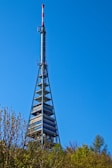 A tall broadcast tower with a red and white antenna at the top against a clear blue sky. The structure features metal frameworks and platforms with satellite dishes visible near the bottom. Surrounding the base of the tower are lush green trees and foliage.