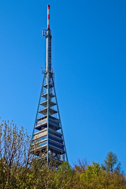 A tall broadcast tower with a red and white antenna at the top against a clear blue sky. The structure features metal frameworks and platforms with satellite dishes visible near the bottom. Surrounding the base of the tower are lush green trees and foliage.