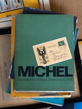 A stack of books with the top one being a green book titled 'MICHEL Ganzsachen-Katalog Deutschland 1980'. On top of the book is an old postcard with some writing and a design featuring what looks like an eagle or a heraldic symbol.