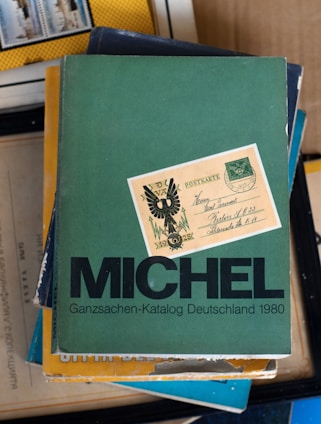 A stack of books with the top one being a green book titled 'MICHEL Ganzsachen-Katalog Deutschland 1980'. On top of the book is an old postcard with some writing and a design featuring what looks like an eagle or a heraldic symbol.