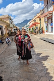 Two women walk down a picturesque cobblestone street lined with colorful buildings and outdoor seating. They hold vibrant woven textiles and appear engaged in conversation. In the background, a person on a wheelchair and another individual are present against a backdrop of mountains and a bright, partly cloudy sky.