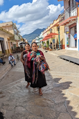 Two women walk down a picturesque cobblestone street lined with colorful buildings and outdoor seating. They hold vibrant woven textiles and appear engaged in conversation. In the background, a person on a wheelchair and another individual are present against a backdrop of mountains and a bright, partly cloudy sky.