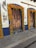Two wooden doors are framed with vibrant orange marigold garlands and green leaves. A sign on the wall reads Los Milagros Café. The building is painted white with blue trim and signs, including one indicating safe travel practices. The stone pavement in front of the doors adds a rustic charm.