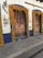 Two wooden doors are framed with vibrant orange marigold garlands and green leaves. A sign on the wall reads Los Milagros Café. The building is painted white with blue trim and signs, including one indicating safe travel practices. The stone pavement in front of the doors adds a rustic charm.