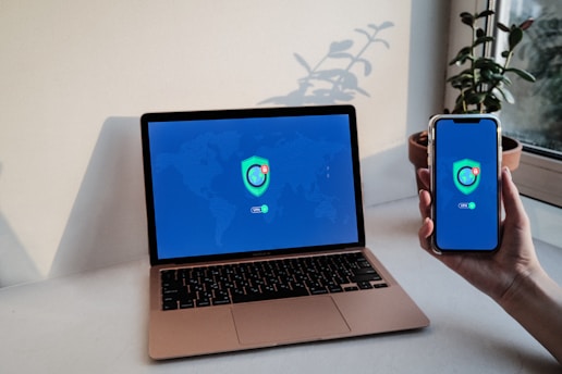 A laptop and a smartphone displaying a VPN login screen with a globe and shield logo. The devices are placed on a white table near a window with a potted plant in the background.