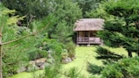 A serene Japanese-style garden with a traditional wooden hut featuring a thatched roof. The garden is lush with various green plants and trees, arranged around rocks. A still pond with green water completes the tranquil setting.