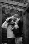 A black and white image depicts a joyful family moment with a man holding a child on his shoulders while a woman kisses the child. They are surrounded by a wooden structure and trees in the background, indicating an outdoor setting.
