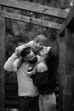 A black and white image depicts a joyful family moment with a man holding a child on his shoulders while a woman kisses the child. They are surrounded by a wooden structure and trees in the background, indicating an outdoor setting.