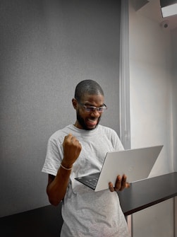 A person with short hair, wearing glasses and a light gray t-shirt, is looking at a laptop with excitement. They have one arm raised in a triumphant gesture. The background is neutral with a textured wall and a soft light from the side.
