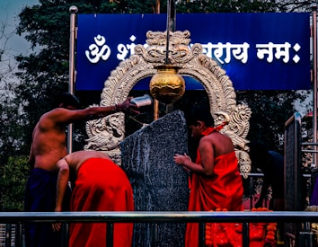 Two people dressed in traditional attire perform a ritual around a large black stone and an intricately designed brass ornament. Water is poured from a metal container over the stone, with a decorated arch in the background, and a blue board displaying text in a script.