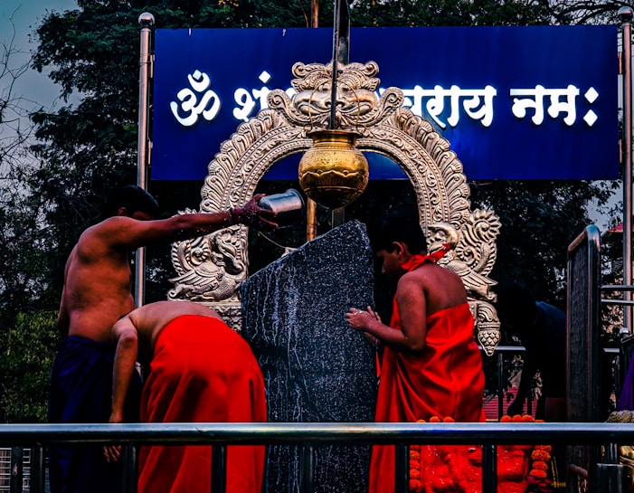 Two people dressed in traditional attire perform a ritual around a large black stone and an intricately designed brass ornament. Water is poured from a metal container over the stone, with a decorated arch in the background, and a blue board displaying text in a script.