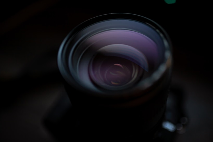 A close-up of a camera lens with reflections visible on its curved glass surface. The lighting is low, enhancing the contrast and creating a moody atmosphere. Subtle reflections and tones of purple and black dominate the image, emphasizing the lens's circular design and mechanical details.