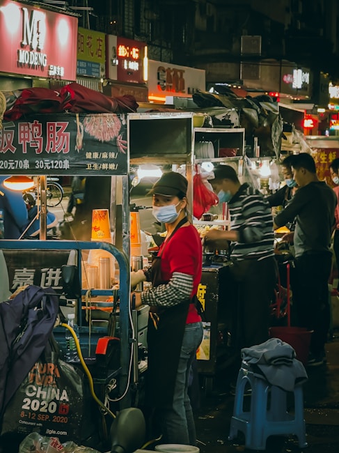 A bustling street food market scene with several food stalls lined up. Vendors are busy preparing food, with one vendor wearing a red shirt and a face mask standing at the forefront. The ambiance is lively with colorful signage and lights illuminating the area.