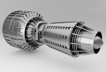 A detailed and intricate metallic structure resembling a jet engine or turbine, featuring various components like fins, vents, and a cylindrical body. The surface has a polished, silver appearance, highlighting the precision of its engineering.