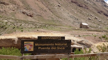 A wooden sign with the text 'Bienvenidos' and 'Monumento Natural Puente del Inca' stands in a rugged, mountainous landscape. In the background, there is a small stone building with a sloped roof, surrounded by sparse vegetation and rocky terrain.