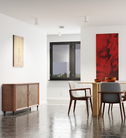 A modern interior space featuring a dining area with a light wooden table and two gray upholstered chairs. The floor is made of reflective dark tiles. On one wall, there is a wooden cabinet with a herringbone pattern, and above it hangs an artwork with a soft beige tone depicting a tower. Adjacent to the table on another wall, a bold red and black abstract painting adds a striking contrast. The room is illuminated by natural light coming through a large window with closed blinds.